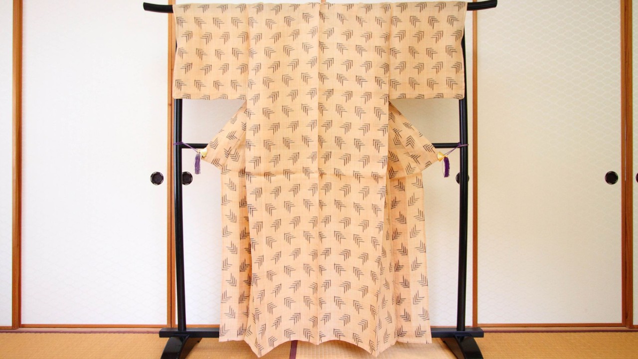 Photograph of a tan kimono suspended on a dark, wooden rack standing on tatami mats, in front of fusuma room dividers.