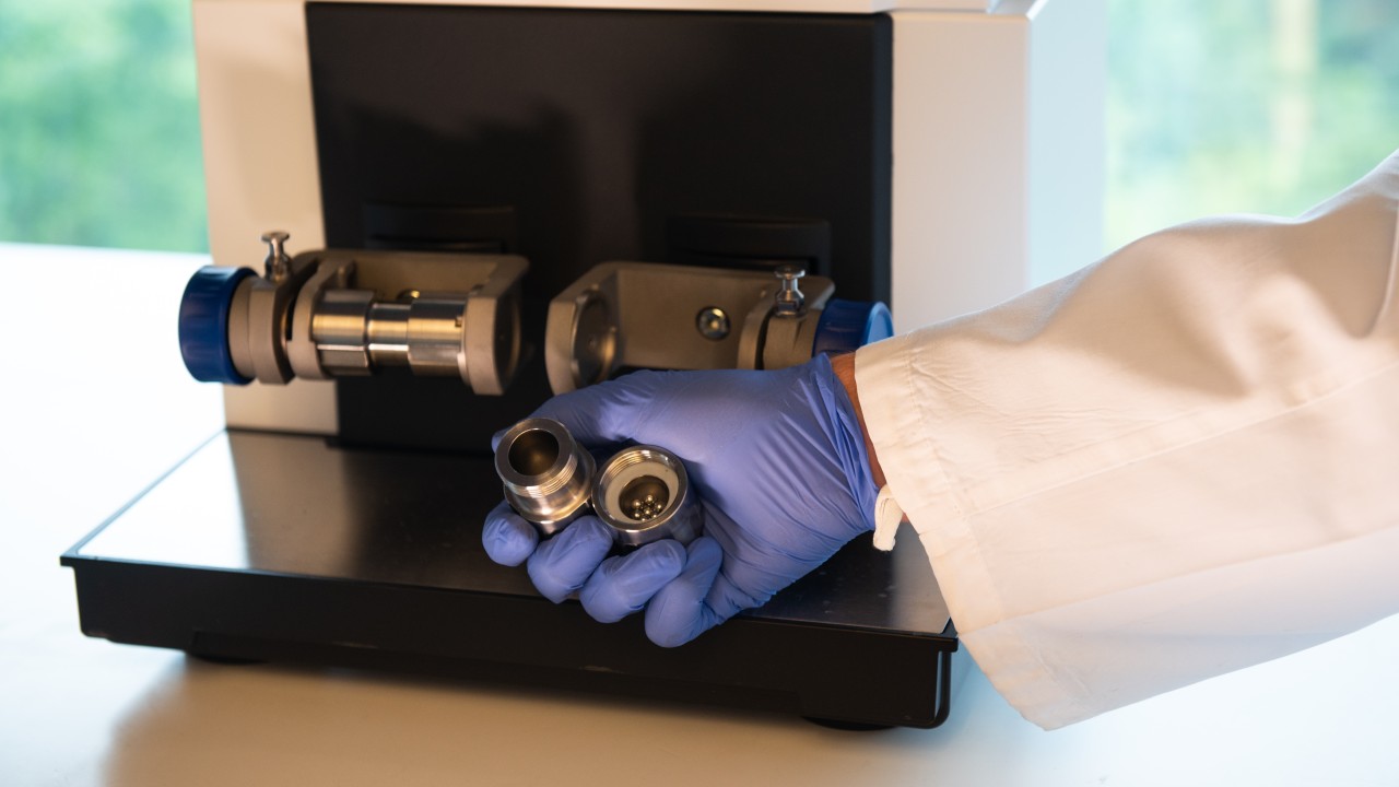 A person in PPE holds a ball milling container, filled with steel balls, in from of the ball-milling machine.