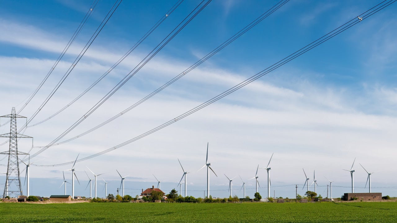 Panoramic photograph of a tall transmission line, with a wind farm in the background against a blue sky.
