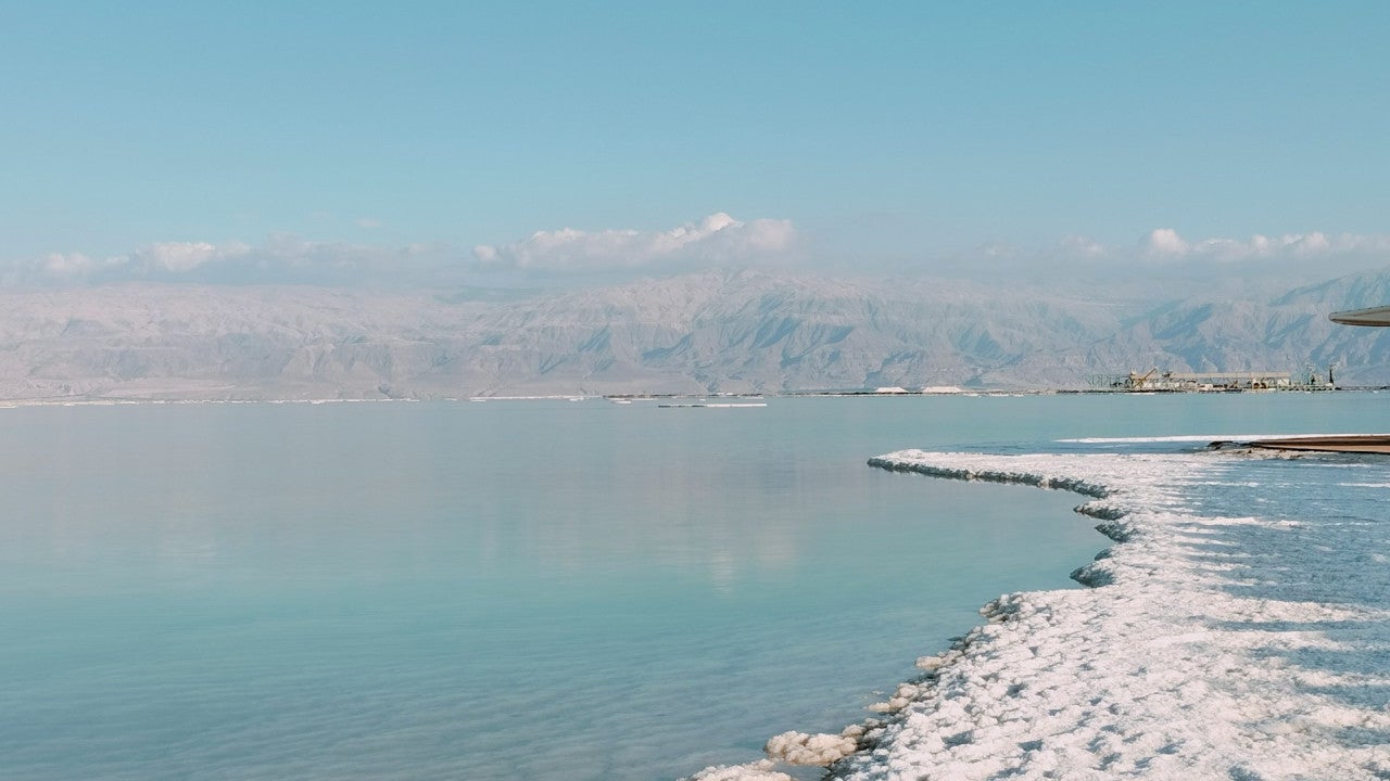 Salt crystals are exposed along the edge of a large, serene body of water. The water reaches some far-off, cloud-capped mountains in the background.