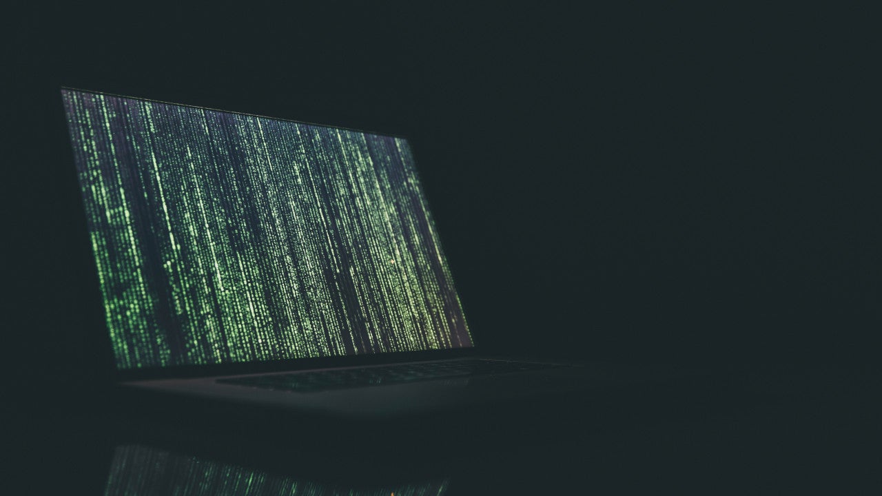 A laptop screen is illuminated against a dark background. The screen is lit up with vertical lines of green code.
