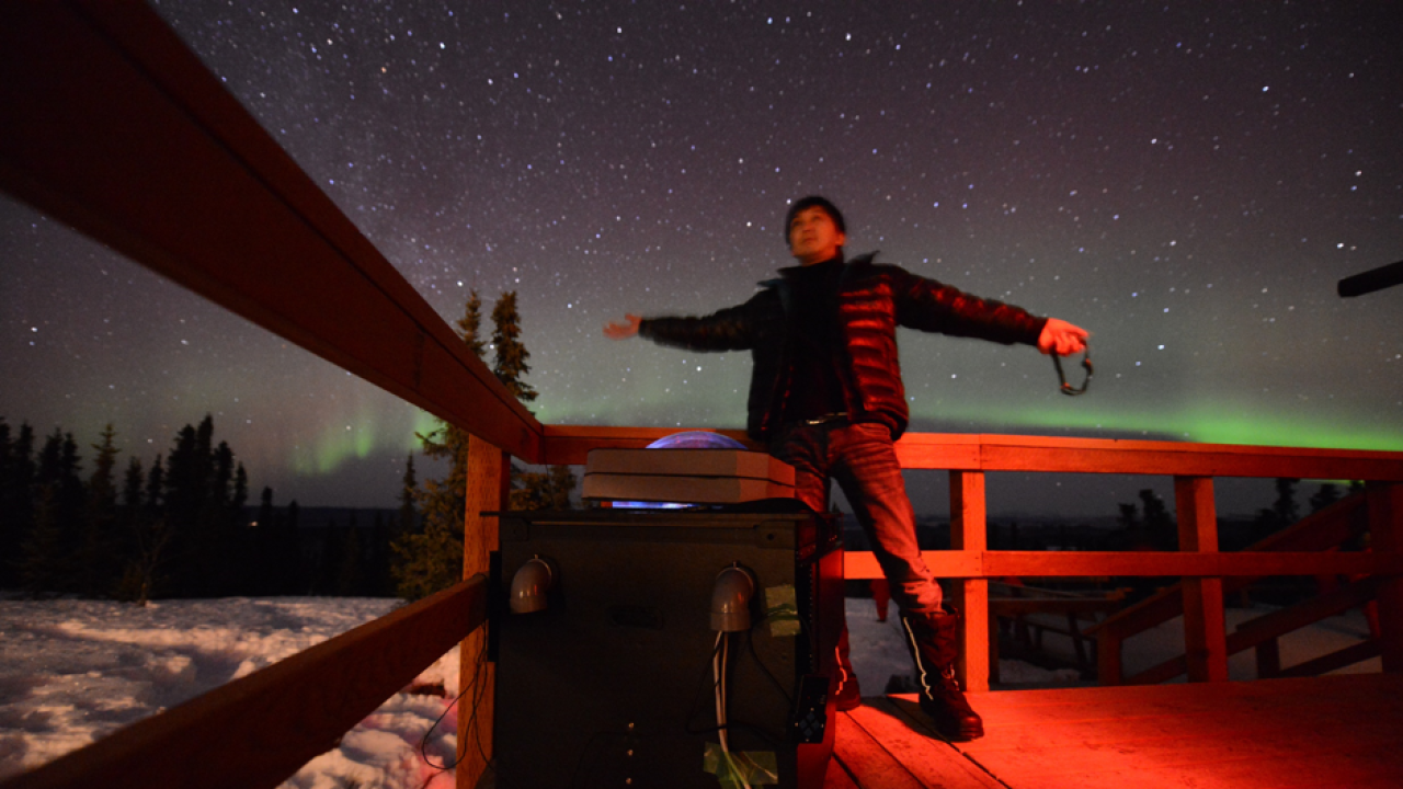 Dr Ryuho Kataoka stands arms spread wide under a bright starry-sky featuring a green aurora. He is next to a fence and some scientific equipment in a snowy tree-lined field. 