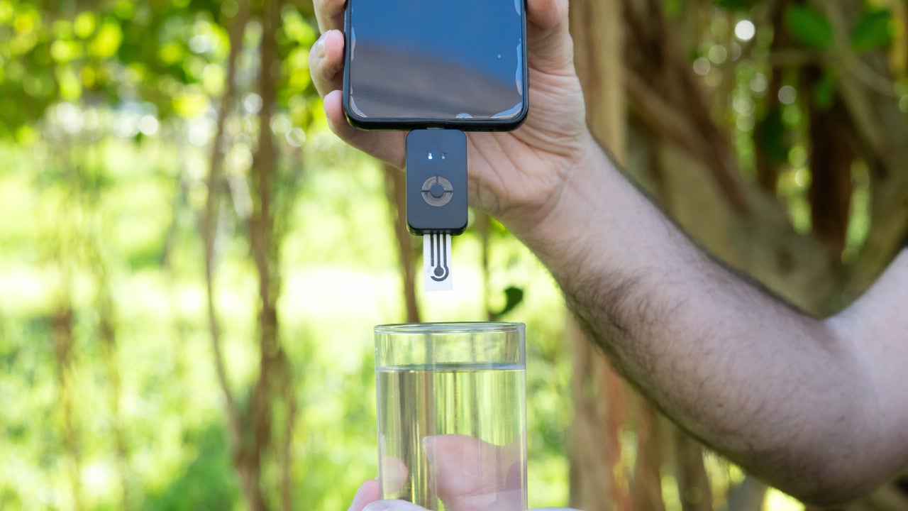 Photograph of a smartphone held above a glass of water against a natural background. A small device is attached to the bottom of the smartphone.