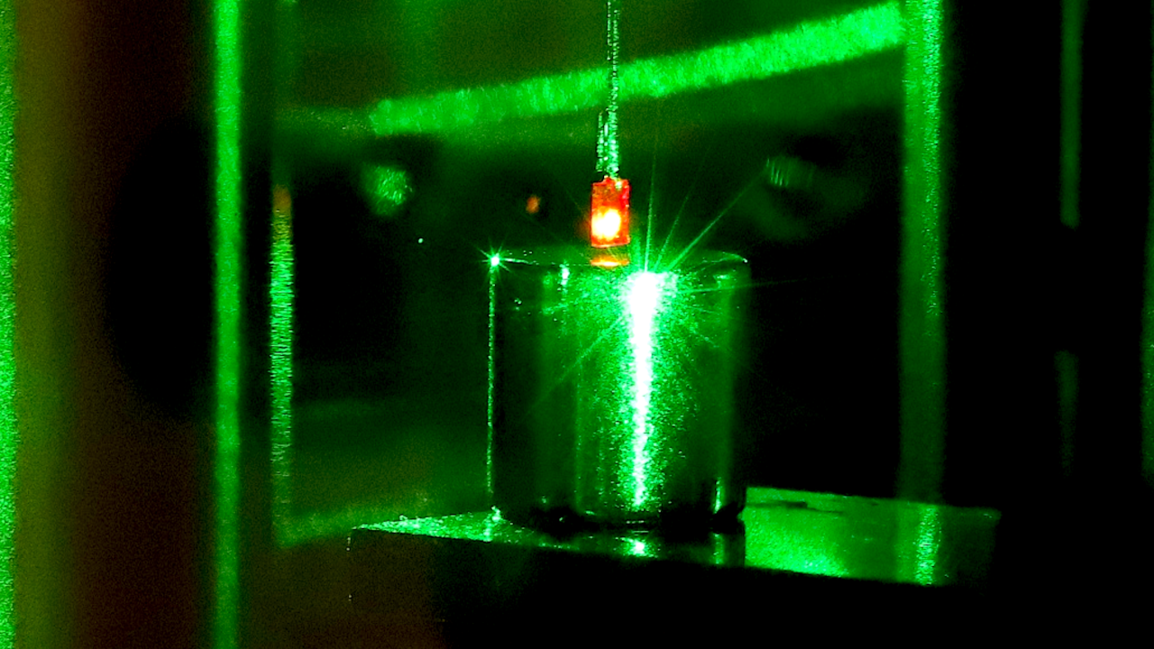 Photograph with a small square diamond glowing red as a result of being illuminated with a green laser, suspended above a smooth cylindrical magnet.