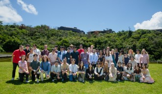 湖の前の芝生に立ち、座る学生たちの集合写真。背後には緑豊かな森とOISTの研究棟が見える。