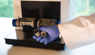 A person in PPE holds a ball milling container, filled with steel balls, in from of the ball-milling machine.
