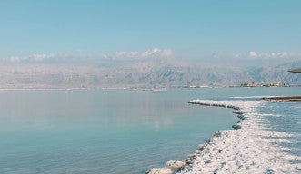 Salt crystals are exposed along the edge of a large, serene body of water. The water reaches some far-off, cloud-capped mountains in the background.