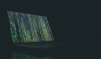 A laptop screen is illuminated against a dark background. The screen is lit up with vertical lines of green code.