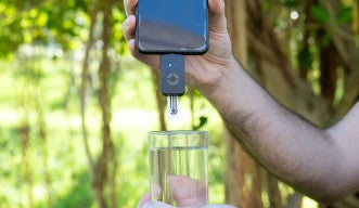 自然を背景に、水の入ったグラスの上にスマートフォンをかざしている。スマートフォンの下部には小型の測定装置が取り付けられている。