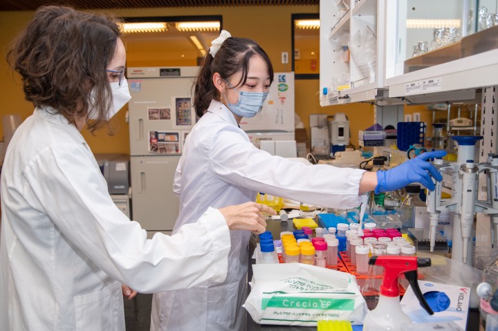Female professor instructing female student in white lab coats at web lab counter with pipettes.