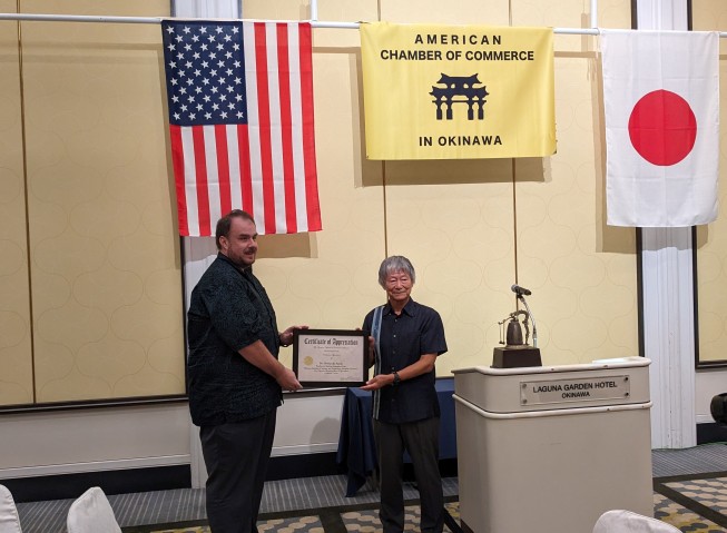 Two men holding a certificate in front of the Japanese, US and American Chamber of Commerce flags