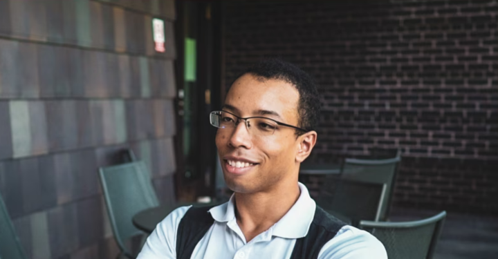 Man with glasses and blue shirt sitting at table with brick wall behind him
