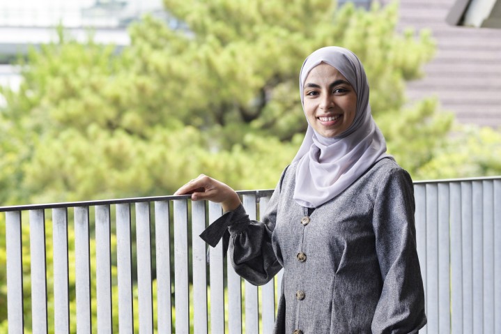 Woman in hijab smiling at camera. Right hand raised on fence with trees and building in background
