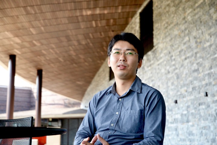 Man with glasses sitting down at table. Stone wall and roof behind and above him.