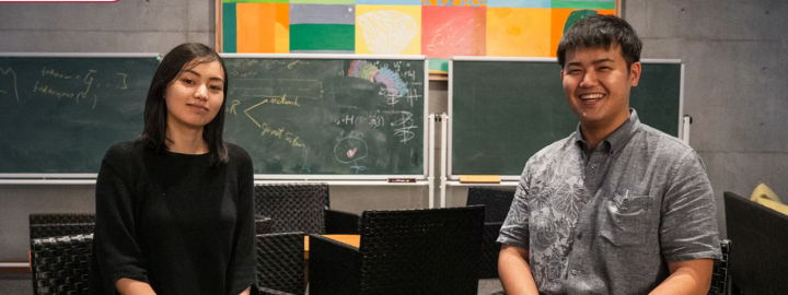 Two students sitting on chairs with chalkboard and black chairs in background