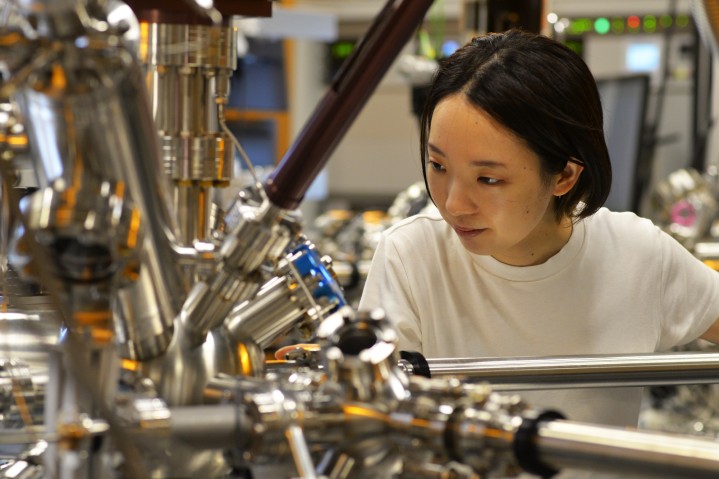 Woman on the right looks down and to the left at a large machine with plenty of pipes