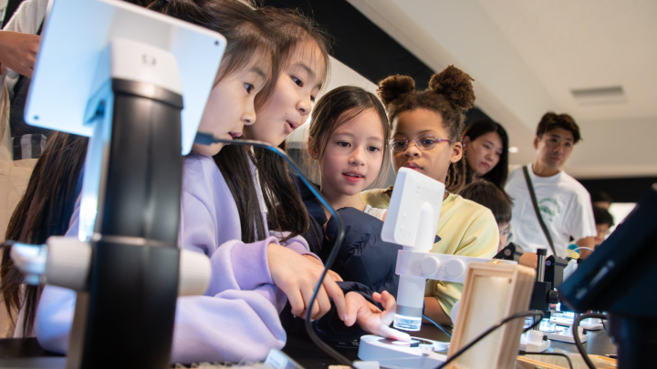 A crowd of children around a digital microscope, their faces fixated on the small display. One of the children has put her finger under the microscope. Adults are watching on in the background. 