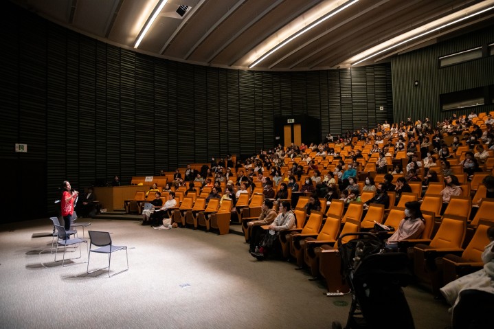 A researcher in a red OIST t-shirt on the stage in front of a mostly full auditorium, dramatically illuminated by the spotlight.