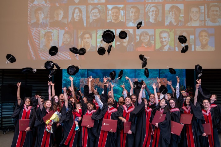 OIST講堂で、アカデミックローブと呼ばれる式服を身にまとった、笑顔の博士号取得者たちが、頭上に帽子を投げている。