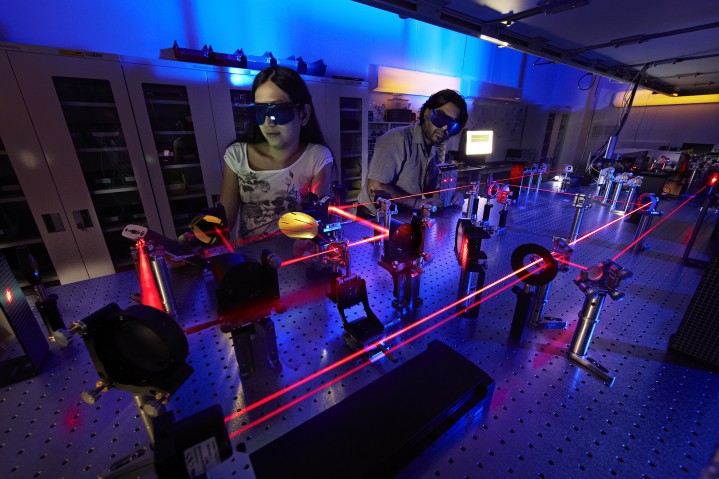 two students wearing dark sunglasses in blue lighting lab. Red lasers bouncing around on mirrors in front of them.