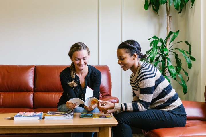Two women sitting on leather couches looking at book in front of them on table.