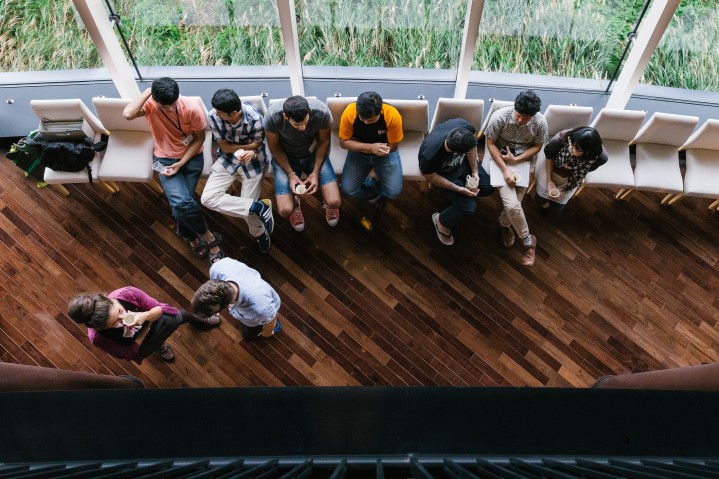 View looking down on floor below in cafe. Seven people sitting in chairs and two standing chatting. Most drinking from cups.