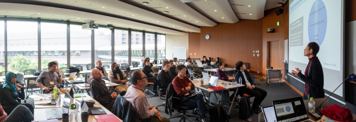 A large group of people sit at desks facing the screen on the right. A man stands in front of the screen, talking.