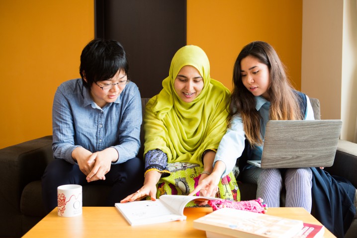 three people sitting on a sofa. Right one has a laptop on their lap and is pointing to a book on a desk in front of them.