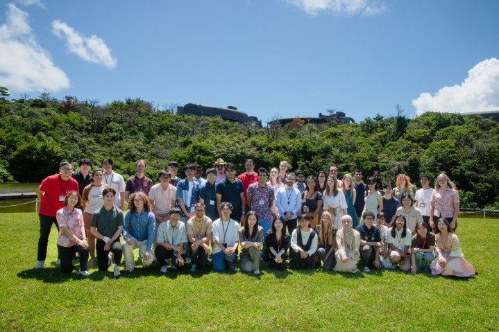 湖の前の芝生に立ち、座る学生たちの集合写真。背後には緑豊かな森とOISTの研究棟が見える。