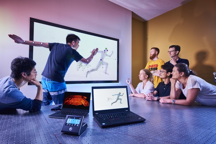 5 people standing and sitting on the right looking at screen in center. Screen has person's model in running motion. Two people on left, one doing running motion. Two laptops in foreground.