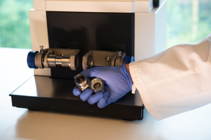 A person in PPE holds a ball milling container, filled with steel balls, in from of the ball-milling machine.