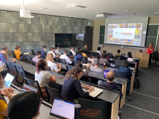 A speaker presenting in front of a screen to an audience seated in a lecture room