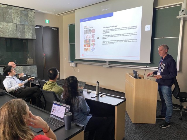 A speaker presenting in front of a screen to an audience seated in a lecture room