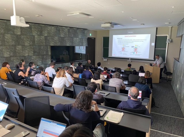 A speaker presenting in front of a screen to an audience seated in a lecture room