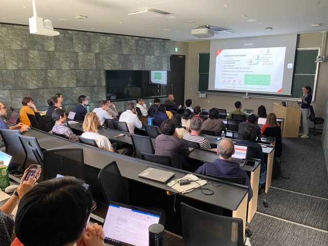 A speaker presenting in front of a screen to an audience seated in a lecture room