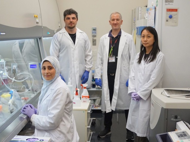 Four people wearing white lab coats standing beside research equipment