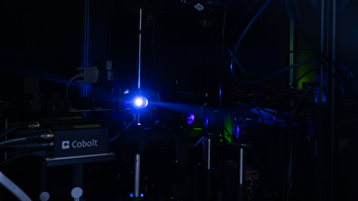 A photograph of a blue laser light hitting a reflective surface inside a complicated spectroscopy setup, housed in a dark area.