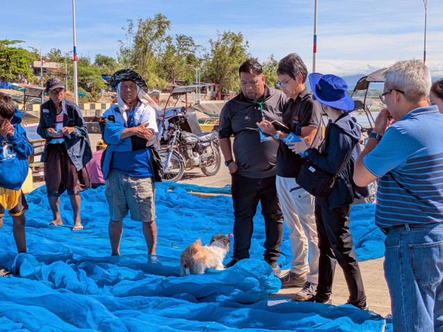 Photograph of a group of fishermen around a researcher, who's noting down answers to questions. They're standing in front of blue fishing nets.