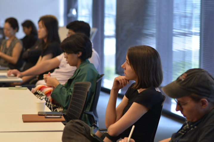 Young people seated in front of multiple tables arranged in a single row.