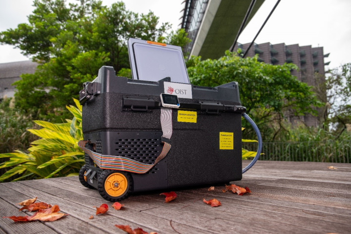 A black box with wheels sits on a deck in front of lush forest. Inside the box resides a 2-chamber prototype egg incubator for aquaculture. On the box, a monitor displays electronic information and the OIST logo is displayed.