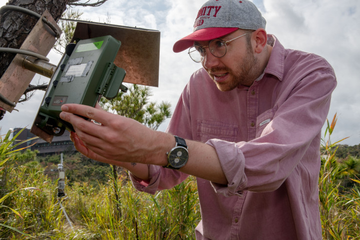 Up close photograph of a man working on a small, bird-house-like scientific device attached to a tree in an overgrown field.