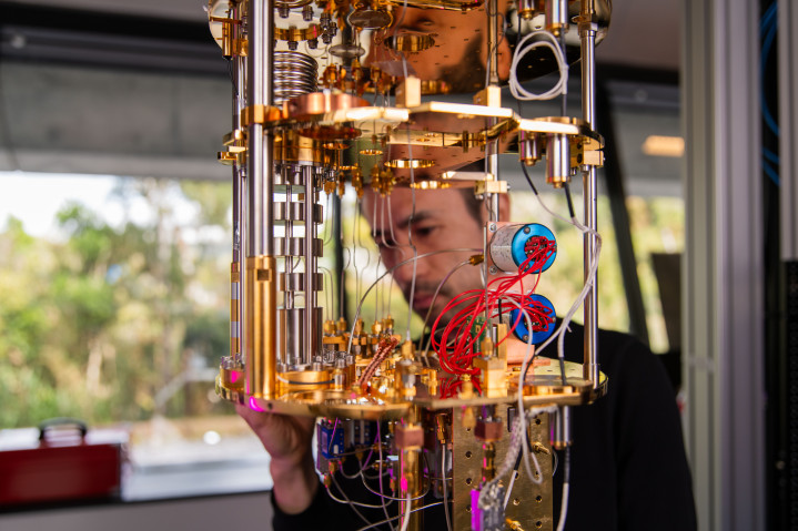 A beautiful gold machine with lots of wires in the foreground, at which a man in the background stares intently. 