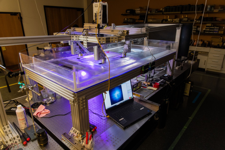 A tank built on top of a lab desk, with a shallow layer of water inside. The tank is lit up from underneath, with a high-speed camera mounted above.