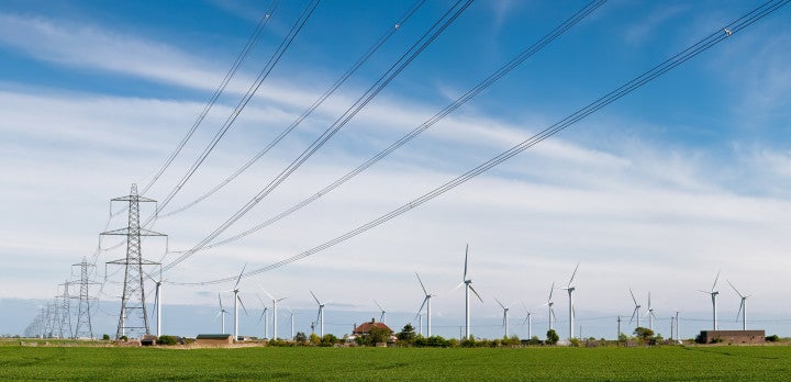Panoramic photograph of a tall transmission line, with a wind farm in the background against a blue sky.