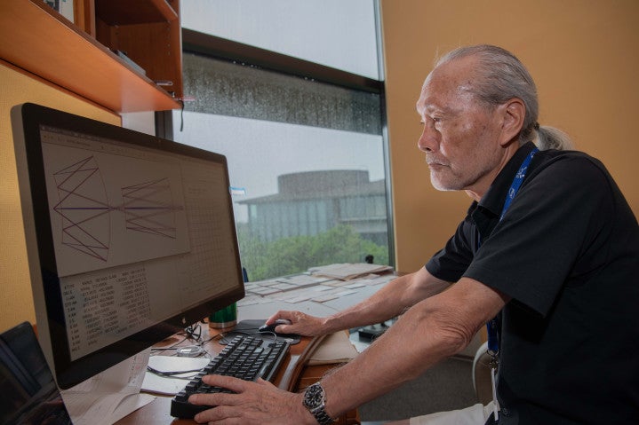 A scientist sits at a desk looking at a computer screen. The screen contains a schematic of a projector design for high NA EUV lithography.