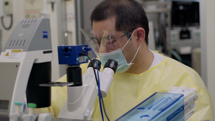 Man in full protective equipment trying to focus a microscope, sitting in the middle of a biology lab.