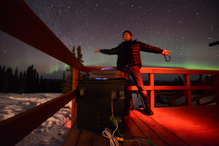 Dr Ryuho Kataoka stands arms spread wide under a bright starry-sky featuring a green aurora. He is next to a fence and some scientific equipment in a snowy tree-lined field. 