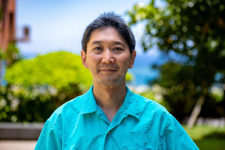 Man in bright blue shirt stands in front of beautiful greenery and ocean.