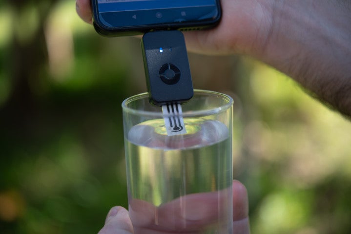 Close-up photograph of a screen-printed electrode attached to a smartphone device being dipped in a glass of water against a lush background. 