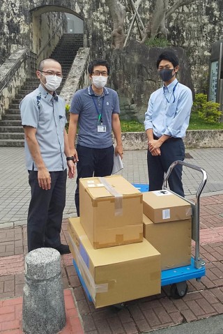 Three men standing next to boxes at the Okinawa Prefectural Government Office.